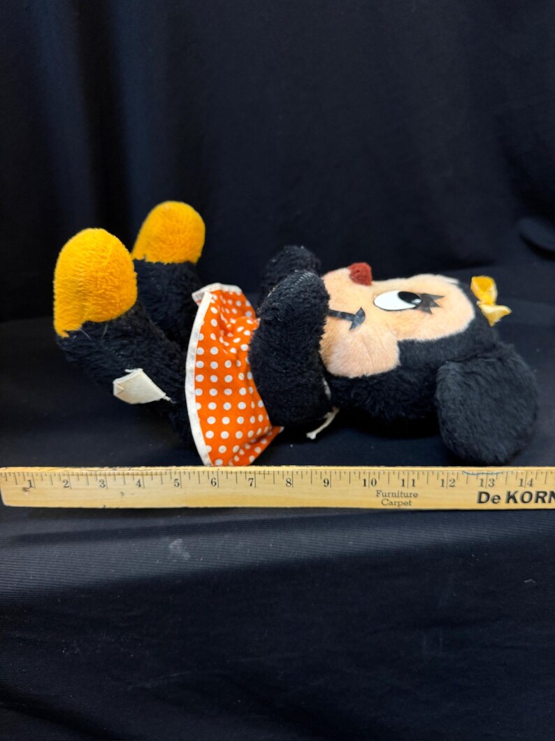 Puede incluir: Un peluche vintage de Minnie Mouse, tumbado boca arriba. El peluche tiene pelo negro, pies amarillos y un vestido de lunares rojos y blancos. Se coloca una regla de madera junto al juguete para la escala.