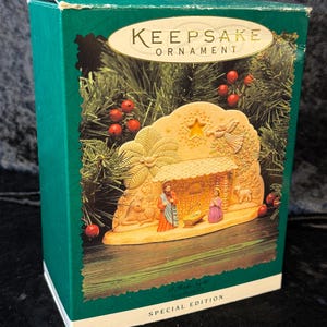 May include: A Christmas ornament in a green box. The ornament depicts a nativity scene with figures and a star. The box has the words "Keepsake Ornament" and "Special Edition" printed on it. The background shows greenery and red berries.
