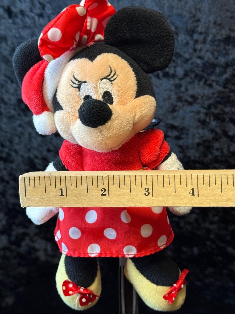 May include: A Minnie Mouse plush doll wearing a red dress with white polka dots, a matching bow, and yellow shoes with red bows. The doll has black ears and a black nose. The doll is approximately 4 inches tall.