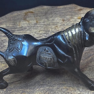 May include: A dark bronze bull figurine with intricate detailing. The sculpture features etched floral and geometric patterns on its body and head. The bull has a stylized mane and horns, and is posed in a walking stance.