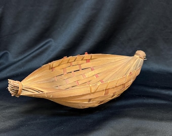 May include: A woven basket made of natural-colored reeds, with a boat-like shape. The basket has a tightly woven base and sides, with red accents. The ends are bundled and tied. The basket is set against a dark background.