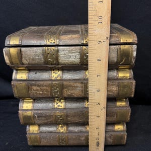 Puede incluir: Una pila de cinco cajas de madera con detalles de lat&oacute;n, que se asemejan a libros antiguos. Las cajas son marrones con adornos y detalles de esquina de metal dorado. Una regla de madera mide aproximadamente 25 cm de altura.