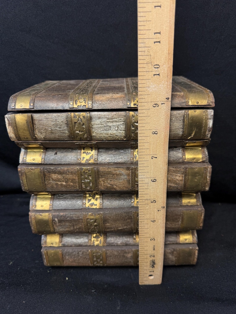 Puede incluir: Una pila de cinco cajas de madera con detalles de lat&oacute;n, que se asemejan a libros antiguos. Las cajas son marrones con adornos y detalles de esquina de metal dorado. Una regla de madera mide aproximadamente 25 cm de altura.