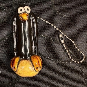 May include: A black and tan penguin-shaped keyring. The penguin has white eyes, an orange beak, and orange feet. A silver ball chain is attached to the top of the penguin.