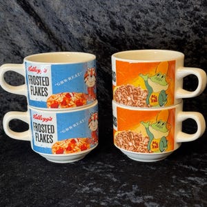 May include: Four white ceramic mugs with handles. Two mugs feature the Kellogg's Frosted Flakes logo with Tony the Tiger and the phrase "GR-R-REAT!". The other two mugs display a cartoon frog with the text "Big Top".