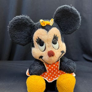 Puede incluir: Peluche vintage de Minnie Mouse. El peluche presenta orejas negras, una cara color melocot&oacute;n y una nariz roja. Lleva un vestido de lunares rojos y blancos y un lazo amarillo. El juguete tiene pies amarillos y est&aacute; sentado.