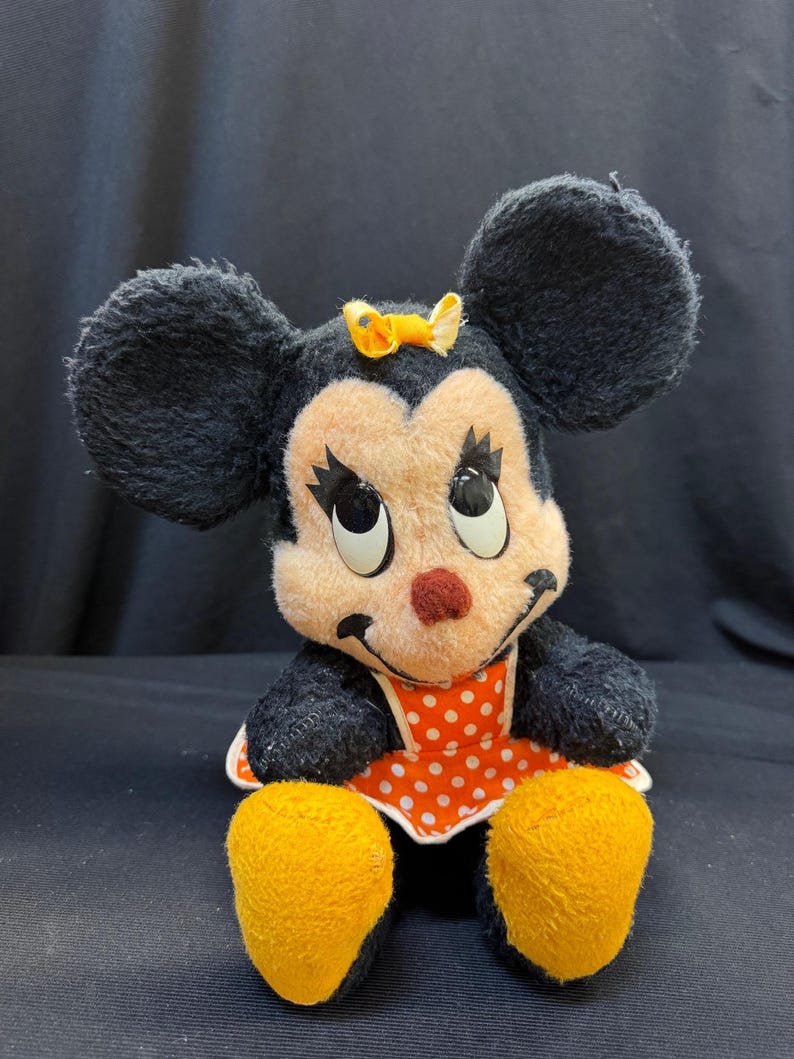 Puede incluir: Peluche vintage de Minnie Mouse. El peluche presenta orejas negras, una cara color melocot&oacute;n y una nariz roja. Lleva un vestido de lunares rojos y blancos y un lazo amarillo. El juguete tiene pies amarillos y est&aacute; sentado.