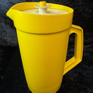 May include: A vibrant yellow plastic jug with a white lid and a handle. The jug features a pouring spout. The lid has a small, yellow knob. The jug is likely designed for serving cold drinks, such as water or juice.