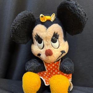Puede incluir: Peluche vintage de Minnie Mouse. El peluche presenta orejas negras, un lazo amarillo y un vestido naranja con lunares blancos. El juguete tiene pies amarillos y una nariz roja. Los ojos son grandes y redondos.