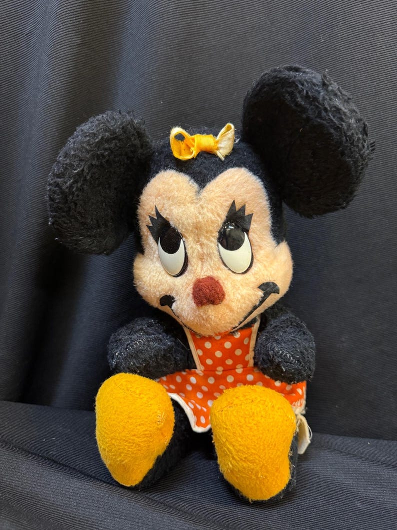 Puede incluir: Peluche vintage de Minnie Mouse. El peluche presenta orejas negras, un lazo amarillo y un vestido naranja con lunares blancos. El juguete tiene pies amarillos y una nariz roja. Los ojos son grandes y redondos.
