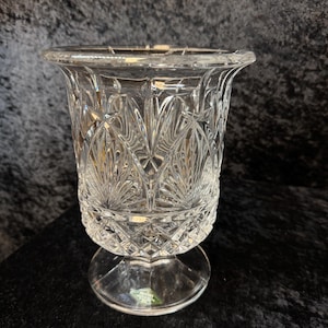 May include: A clear, cut-glass vase with a flared rim and a pedestal base. The vase features a detailed, repeating pattern of diamond and fan shapes. The glass is transparent, allowing light to pass through and highlight the intricate design.