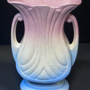 May include: A vintage ceramic vase with a two-handled design. The vase features a color gradient, transitioning from a light pink at the top to white in the middle, and ending with a light blue base. The vase has a scalloped rim and measures approximately 8 inches tall.