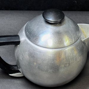 May include: A vintage aluminum teapot with a black handle and lid knob. The teapot has a rounded body and a spout for pouring. The handle is curved and attached to the side of the pot. The lid knob is round and black.