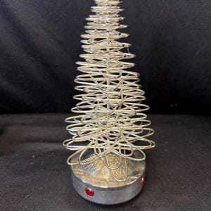 Op de afbeelding: Een zilveren metalen kerstboomdecoratie. De boom is gemaakt van opgerold draad en heeft een conische vorm. De basis is een zilveren cilinder met drie rode juweelaccenten. De boom is ongeveer 20 cm hoog.