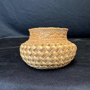 May include: A small, round woven basket with a flared rim. The basket is made of natural-coloured reeds and has a textured, patterned design. The basket is likely handmade and suitable for storage or decoration.