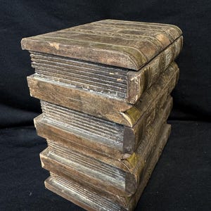 Puede incluir: Una caja decorativa con forma de pila de libros. La caja es de madera con un aspecto envejecido y desgastado. Los bordes est&aacute;n acentuados con metal color lat&oacute;n. Una soluci&oacute;n de almacenamiento &uacute;nica.