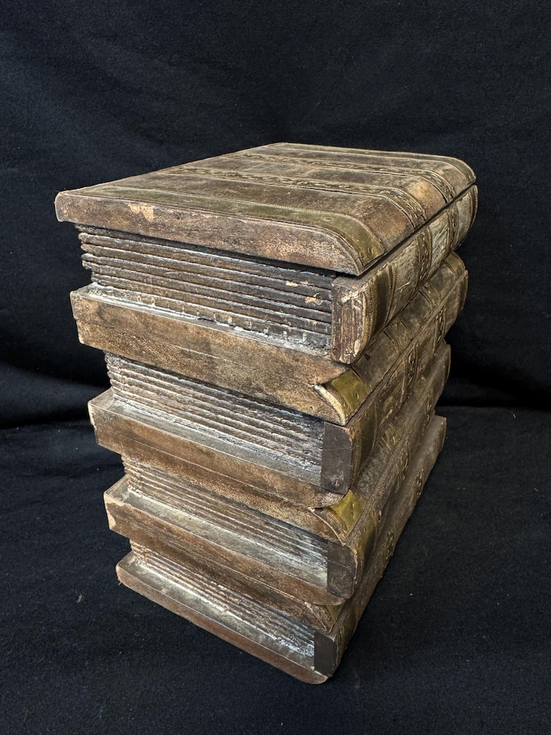 Puede incluir: Una caja decorativa con forma de pila de libros. La caja es de madera con un aspecto envejecido y desgastado. Los bordes est&aacute;n acentuados con metal color lat&oacute;n. Una soluci&oacute;n de almacenamiento &uacute;nica.