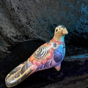 Puede incluir: Figurilla de pájaro de cerámica pintada a mano. El pájaro tiene una cabeza de color canela, un collar rojo y un cuerpo adornado con patrones florales rosas, negros y azules. El pájaro está sobre una superficie negra.