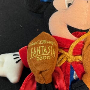 K&ouml;nnte beinhalten: Ein Mickey Mouse Pl&uuml;schtier, das als Zauberer aus Disneys Fantasia verkleidet ist. Die Puppe tr&auml;gt einen blauen Hut, eine rote Robe und braune Handschuhe. Der Handschuh tr&auml;gt den Text "Walt Disney Fantasia 2000" in Gold.