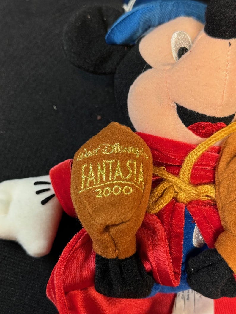 K&ouml;nnte beinhalten: Ein Mickey Mouse Pl&uuml;schtier, das als Zauberer aus Disneys Fantasia verkleidet ist. Die Puppe tr&auml;gt einen blauen Hut, eine rote Robe und braune Handschuhe. Der Handschuh tr&auml;gt den Text "Walt Disney Fantasia 2000" in Gold.