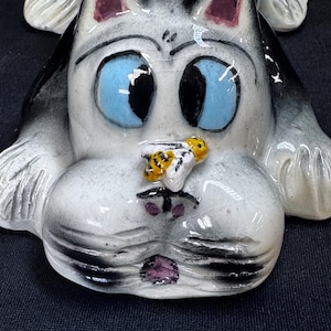 Puede incluir: Figura de gato de cerámica con un diseño caprichoso. El gato tiene grandes ojos azules, pelaje gris y blanco, y una pequeña abeja en la nariz. Las orejas están acentuadas con un color rosa intenso.