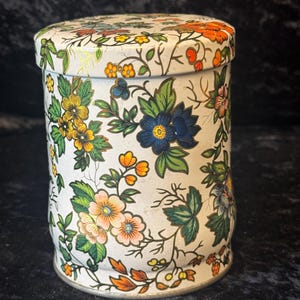 Peut inclure: Une boîte cylindrique avec un couvercle, ornée d'un motif floral. Le design présente une base blanche avec des fleurs colorées dans les tons de bleu, jaune, orange et rose, ainsi que des feuilles vertes et des tiges brunes. La boîte est probablement utilisée pour le stockage.