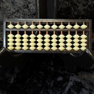May include: A vintage abacus with a dark wooden frame and ivory-colored beads. The abacus is displayed on a black velvet surface, showcasing its traditional design. The beads are arranged on wooden rods, ready for calculations.