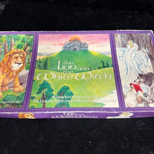 May include: A vintage board game box titled "The Lion, the Witch, and the Wardrobe" with illustrations. The box features a lion, a castle on a hill, and a scene with a wolf and a figure in white. The box is purple with colorful artwork.