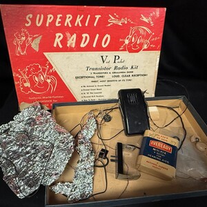 Op de afbeelding: Vintage Superkit Radio transistorradiokit in een rode doos met de tekst "Superkit Radio". De kit bevat een zwarte radio, een batterij en andere componenten. De doos heeft illustraties van stripfiguren.