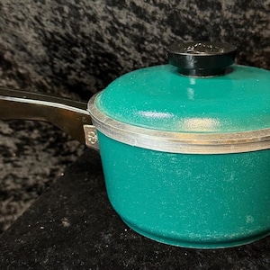 May include: A teal-colored saucepan with a lid and a long, dark handle. The pot has a silver rim and a black knob on the lid. The handle is attached to the pot with a metal bracket. The pot is sitting on a dark surface.