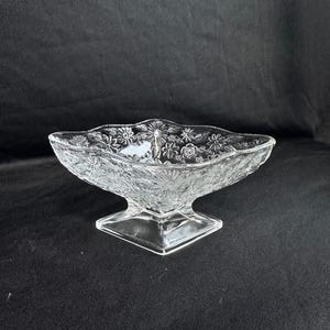 May include: Clear glass serving dish with a floral pattern. The dish has a rectangular base and a diamond-shaped bowl. The bowl is decorated with embossed flowers and leaves. The dish is likely used for serving desserts or snacks.