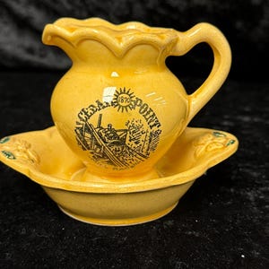 May include: A yellow ceramic miniature pitcher and basin set. The pitcher has a handle and a scalloped rim. The basin has a scalloped edge and green floral accents. The pitcher has a black graphic with the text "Cedar Point" and "1870".