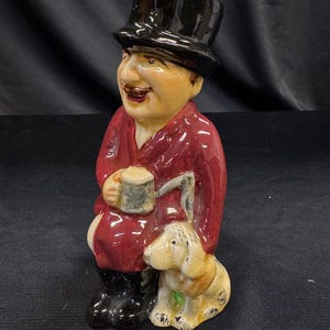 Puede incluir: Figura de cerámica de un hombre con abrigo rojo y sombrero de copa negro, sosteniendo una jarra. Un perro pequeño está sentado a sus pies. La figura tiene un acabado brillante y está pintada con detalles.