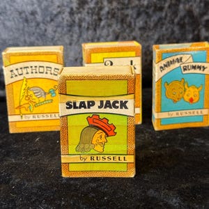 May include: A collection of vintage card games, including "Slap Jack," "Authors," and "Animal Rummy." The boxes are rectangular with colorful illustrations and text. The "Slap Jack" box features a profile of a person wearing a red hat. The games are by Russell.