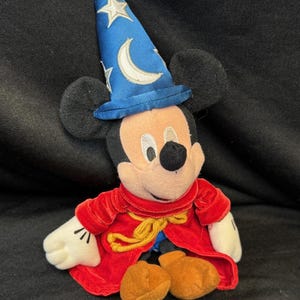 K&ouml;nnte beinhalten: Ein Stofftier von Mickey Mouse als Zauberlehrling aus Fantasia. Mickey tr&auml;gt einen blauen Zauberhut mit Sternen und einem Mond, einen roten Samtumhang und braune Schuhe. Die Disney-Figur sitzt.