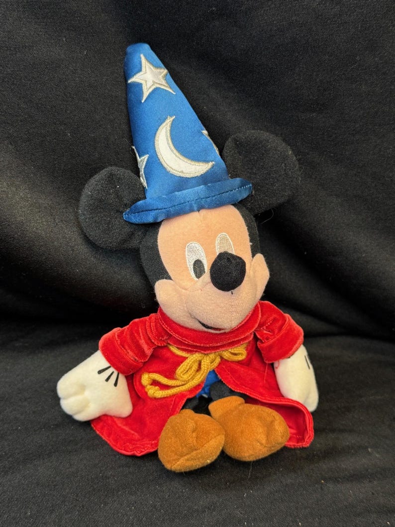 K&ouml;nnte beinhalten: Ein Stofftier von Mickey Mouse als Zauberlehrling aus Fantasia. Mickey tr&auml;gt einen blauen Zauberhut mit Sternen und einem Mond, einen roten Samtumhang und braune Schuhe. Die Disney-Figur sitzt.