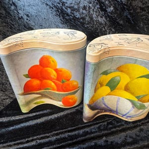 May include: Two decorative canisters with lids. One canister features a painting of oranges in a bowl, the other lemons. The lids are beige with faded illustrations. The canisters are painted with a light blue background.