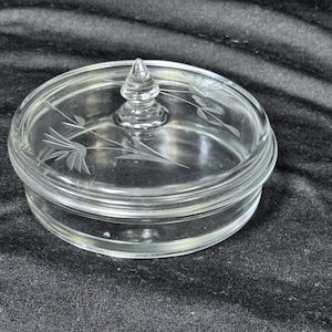May include: A clear glass jar with a lid. The lid has a conical handle and etched floral designs. The jar is round and sits on a dark surface. The jar is made of clear glass.