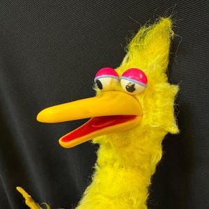 May include: A plush yellow bird puppet with a long yellow beak and red mouth. The bird has large, round eyes with pink and blue accents. The puppet is made of a soft, fuzzy material.