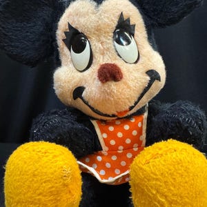 Puede incluir: Juguete de peluche vintage con un personaje de rat&oacute;n sonriente con pelaje negro, orejas grandes y manos amarillas. El rat&oacute;n tiene una cara color melocot&oacute;n, una nariz roja y un lazo amarillo. Lleva un babero naranja con lunares blancos.