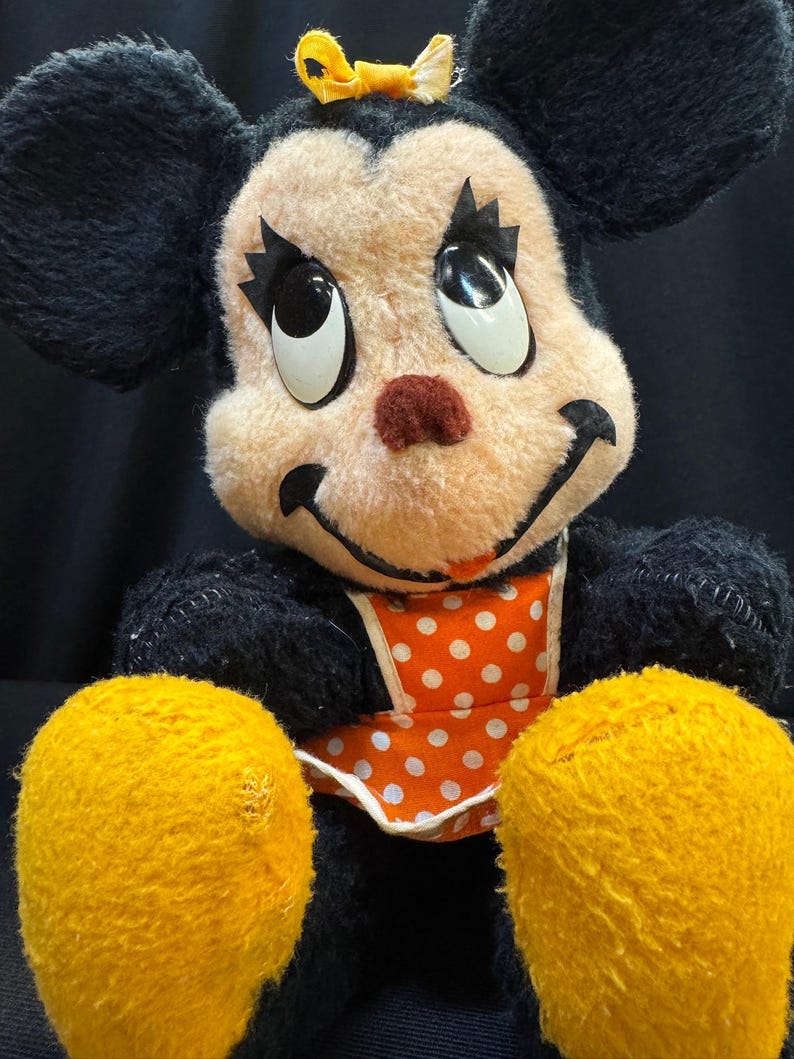 Puede incluir: Juguete de peluche vintage con un personaje de rat&oacute;n sonriente con pelaje negro, orejas grandes y manos amarillas. El rat&oacute;n tiene una cara color melocot&oacute;n, una nariz roja y un lazo amarillo. Lleva un babero naranja con lunares blancos.