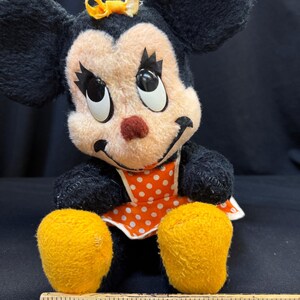 Puede incluir: Mu&ntilde;eca de peluche vintage de Minnie Mouse con pelaje negro, cara color melocot&oacute;n y ojos grandes y expresivos. Lleva un lazo amarillo, una nariz roja y un delantal de lunares naranja y blanco. Se ven sus pies amarillos. Una regla de madera est&aacute; en la parte inferior.