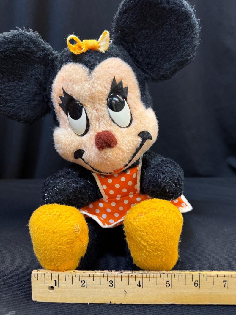 Puede incluir: Mu&ntilde;eca de peluche vintage de Minnie Mouse con pelaje negro, cara color melocot&oacute;n y ojos grandes y expresivos. Lleva un lazo amarillo, una nariz roja y un delantal de lunares naranja y blanco. Se ven sus pies amarillos. Una regla de madera est&aacute; en la parte inferior.