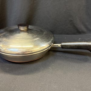 Puede incluir: Una sartén de acero inoxidable con tapa. La sartén tiene un mango negro y un pequeño pomo negro en la tapa. La sartén es redonda y tiene un fondo color cobre.