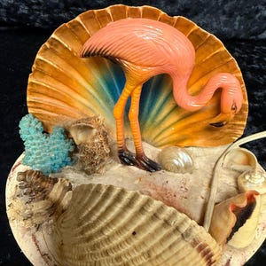 May include: A decorative flamingo figurine with a pink body, orange legs, and black feet. The flamingo stands in front of a large, open seashell with a sandy base, surrounded by smaller shells and coral. The seashell has a gradient of colors.
