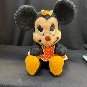 Puede incluir: Un peluche vintage de Minnie Mouse. El peluche presenta pelaje negro, orejas grandes y redondas y una cara sonriente con una nariz roja. Lleva un vestido de lunares rojos y blancos y tiene pies amarillos y un lazo amarillo.