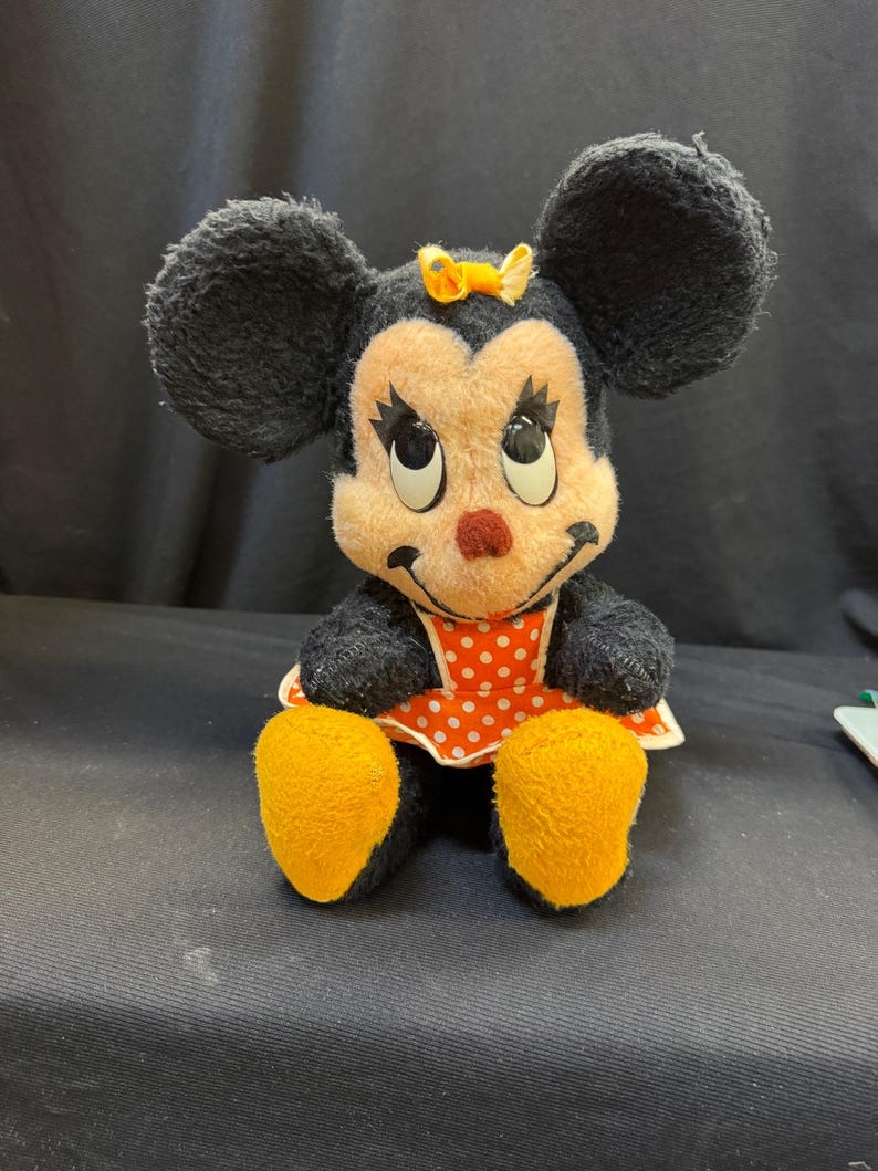 Puede incluir: Un peluche vintage de Minnie Mouse. El peluche presenta pelaje negro, orejas grandes y redondas y una cara sonriente con una nariz roja. Lleva un vestido de lunares rojos y blancos y tiene pies amarillos y un lazo amarillo.