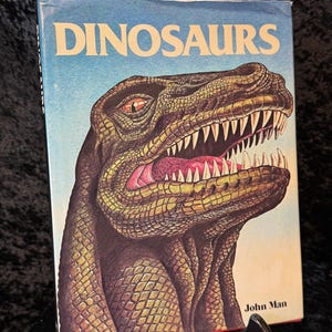 以下が含まれることがあります： 「DINOSAURS」というタイトルのヴィンテージ本。表紙には恐竜の頭部の詳細なイラストが描かれています。恐竜は緑と茶色の鱗、鋭い歯、赤い舌で描かれています。著者の名前、John Man、は下部に印刷されています。
