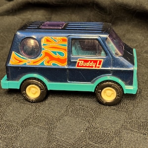 Op de afbeelding: Een vintage Buddy L speelgoedbusje in donkerblauw met teal accenten. De bus heeft een vlammenontwerp in oranje, geel en teal aan de zijkant, en het Buddy L-logo. De wielen zijn beige.
