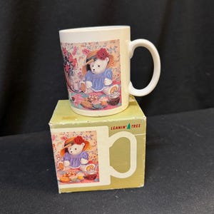 Op de afbeelding: Witte keramische mok met een handvat, met een teddybeer illustratie in een blauwe jurk en een hoed met een rode roos. De mok staat op een bijpassende doos met de tekst "LEANIN' TREE". Het ontwerp bevat bloemen en voedsel elementen.